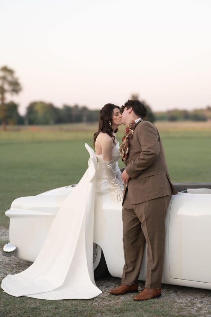 Groom and bride pose for a kiss on a vintage car.